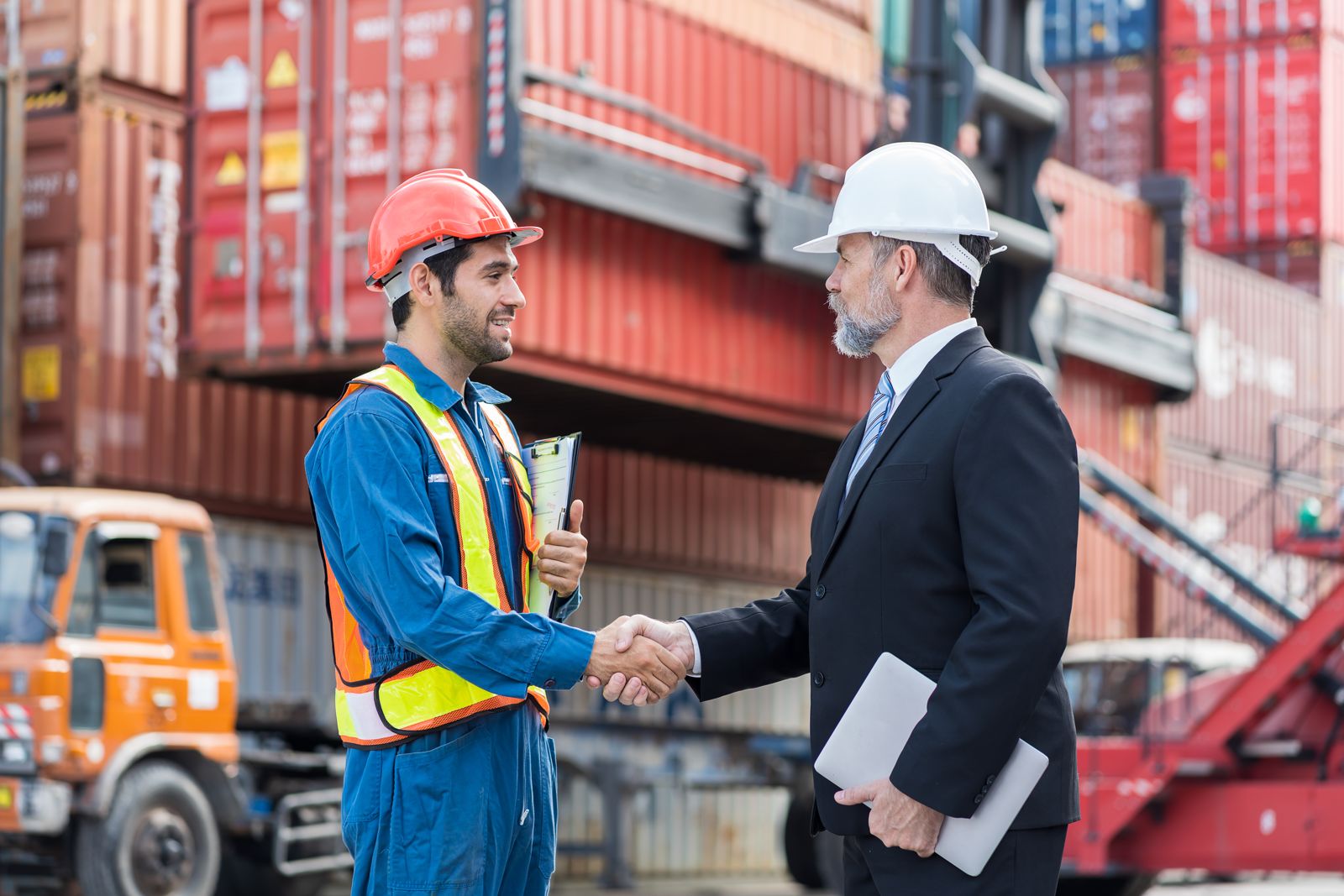 Two men shaking hands in front of stacked shipping containers at a terminal